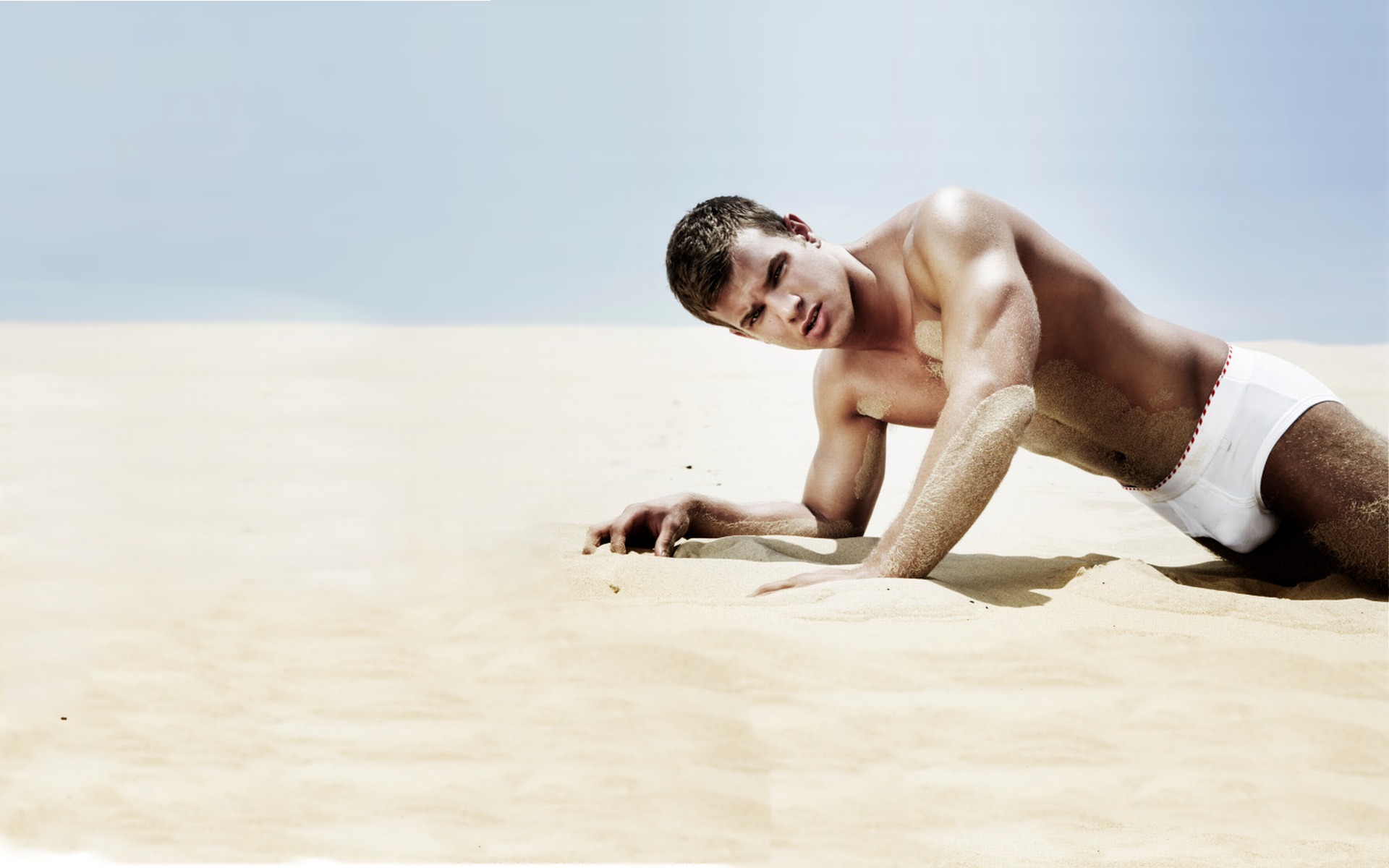 молодой парень в белых плавках ползет по песку, фото красивого мужчины